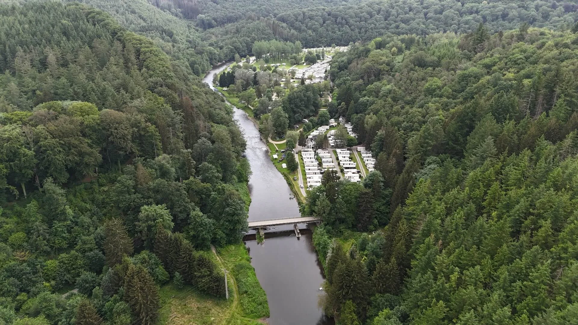 Dronebeeld van een camping met stacaravans langs een rivier, gelegen in een dichtbebost dal met heuvels en brug in het midden van het beeld