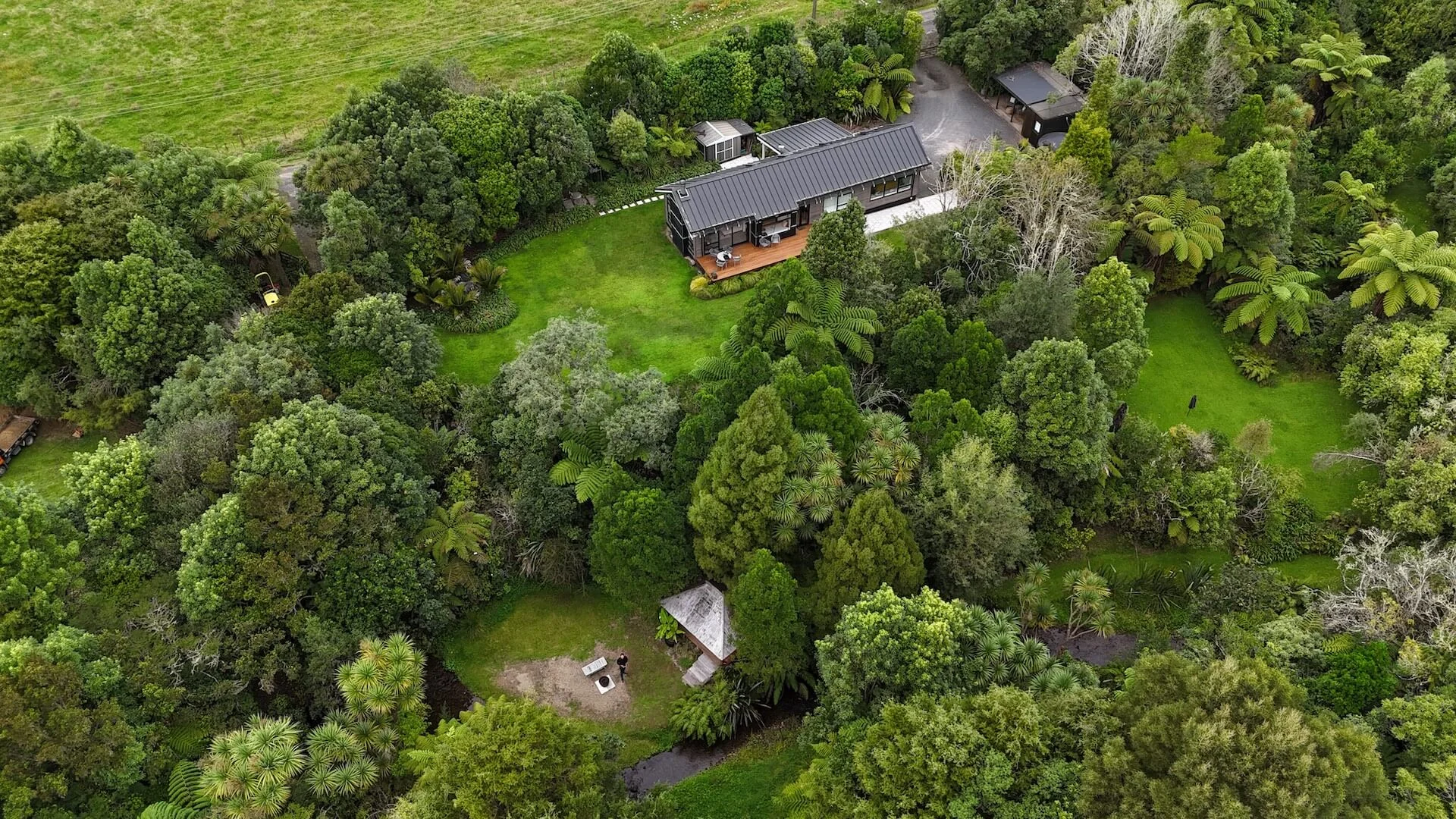 Dronebeeld van een vrijstaande moderne woning met houten terras, omgeven door dichtbegroeid groen en grasvelden in een bosrijke omgeving