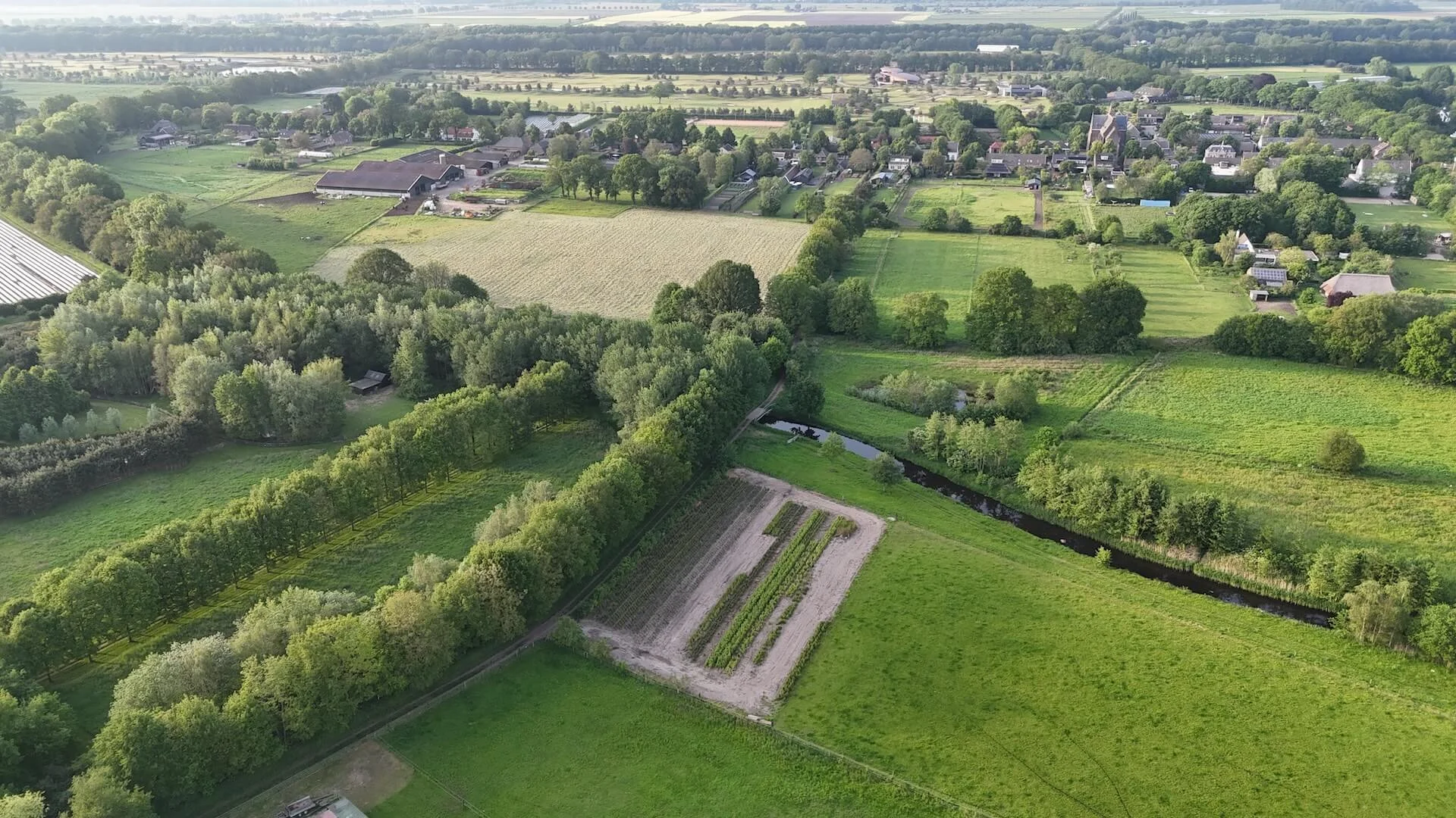 Droneopname van een groen en agrarisch landschap in Nederland met bomen, akkers, een beekje en een dorp op de achtergrond