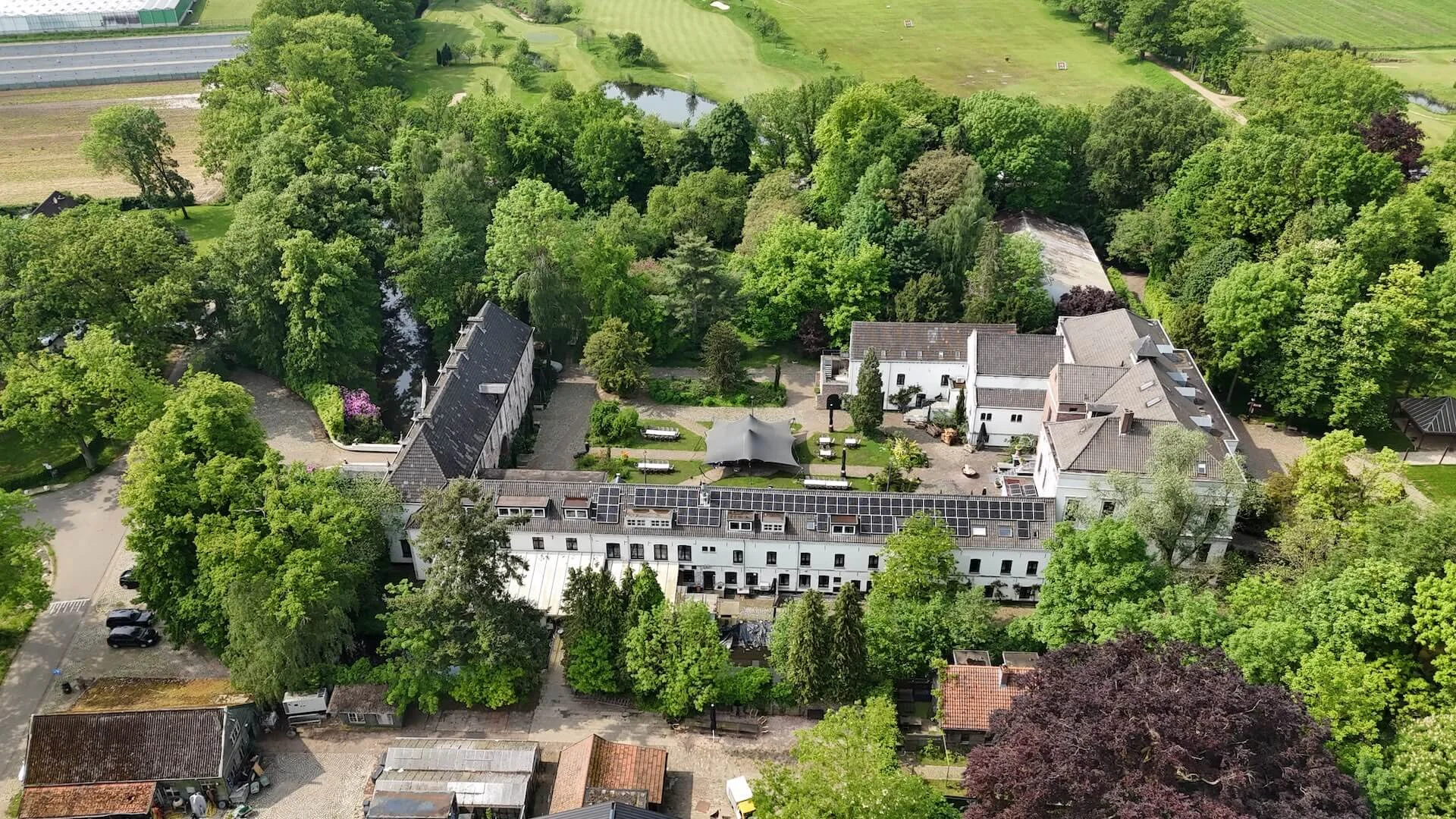 Droneopname van een groot wit landhuis met binnenplaats, gelegen in een bosrijke omgeving, mogelijk gebruikt als trouwlocatie of evenemententerrein